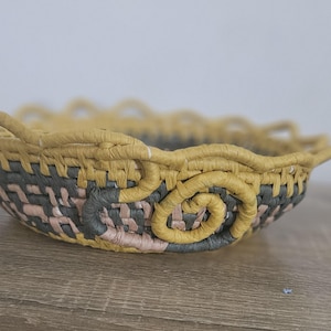 May include: A shallow, round woven basket featuring a scalloped rim in mustard yellow. The body of the basket is constructed from a mix of dark grey, mustard yellow, and soft pink woven fibres, with a prominent spiral design on the exterior side.