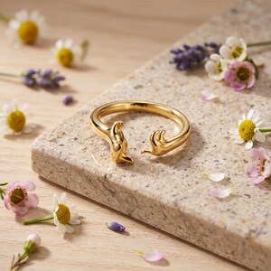 May include: A polished gold-tone open ring featuring two stylized hands reaching toward each other at the center. The band is smooth and circular, with the hand motifs forming the open ends of the ring, creating a continuous metallic loop.