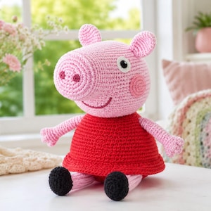Pode incluir: Um peluche artesanal de croché representando uma personagem de porco com uma cabeça grande cor-de-rosa, um focinho proeminente e duas orelhas. A figura usa um vestido vermelho vivo, tem braços e pernas rosa-claro e sapatos pretos.