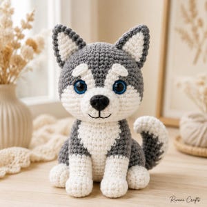 Peut inclure: Un jouet chiot husky crocheté à la main avec un motif en laine grise et blanche, de grands yeux bleus en plastique, un nez noir brodé et une petite queue recourbée. L'animal en peluche est assis avec des oreilles pointues.