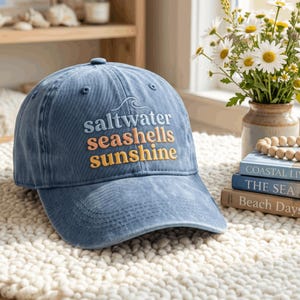 May include: A blue baseball cap with a distressed finish featuring the embroidered text "saltwater" in light blue, "seashells" in peach, and "sunshine" in yellow, topped with a small embroidered wave graphic above the lettering.