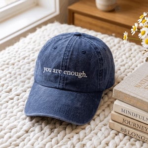 Op de afbeelding: Een marineblauwe honkbalpet met een verweerde, verwassen textuur. Op de voorkant van de pet staat de witte geborduurde tekst "you are enough.". De pet heeft een gestructureerde kroon, een gebogen klep en kleine ventilatiegaatjes bovenop.