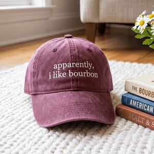 Può includere: Un cappellino da baseball color marrone con finitura vissuta, caratterizzato dalla scritta ricamata in bianco "apparently, i like bourbon" sul pannello frontale. Il cappellino presenta una visiera curva, un bottone superiore e occhielli.
