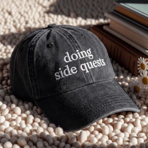 Könnte beinhalten: Eine anthrazitfarbene Baseballkappe mit verwaschenem, abgenutztem Finish. Auf der Vorderseite befindet sich eine weiße Stickerei mit dem Text "doing side quests" in einer klaren Serifenschrift. Die Kappe hat eine strukturierte Krone.
