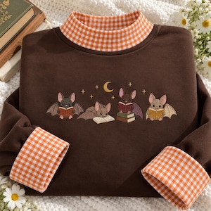 May include: A dark brown long-sleeve sweatshirt featuring an orange and white gingham mock neck collar and matching sleeve cuffs. The chest displays an embroidered graphic of four small bats reading books under a crescent moon and stars.