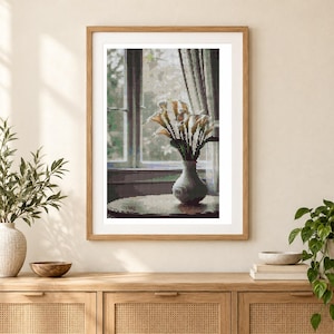May include: A framed wall art print featuring a pixelated image of a bouquet of white calla lilies in a rounded vase. The vase sits on a table in front of a window with soft light filtering through. The frame is a light wood color.