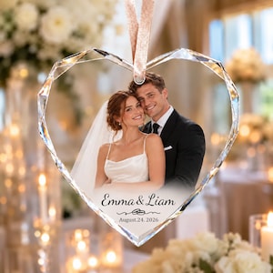 May include: A heart-shaped crystal ornament featuring a printed photograph of a couple in wedding attire. The text reads Emma & Liam with a graphic of two interlocking rings and the date August 24, 2026. A light pink ribbon is attached to the top.