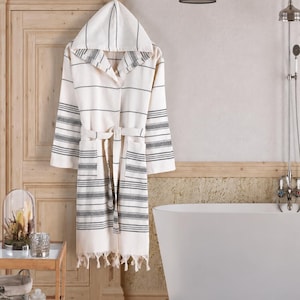 May include: A cream-colored hooded bathrobe featuring horizontal dark gray stripes, long sleeves, a matching tie belt at the waist, two front pockets, and a decorative fringe hemline along the bottom edge of the garment.