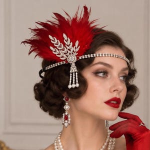 May include: A headpiece featuring vibrant red feathers and a silver-toned metal ornament shaped like a leaf, embellished with clear crystals and small white pearls. The design includes a thin, crystal-encrusted band that encircles the head.