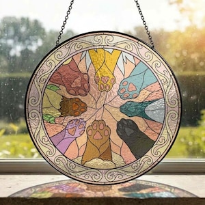 May include: A circular stained glass suncatcher featuring a central design of eight colorful animal paws arranged in a radial pattern. The paws are rendered in shades of brown, yellow, blue, green, and black, enclosed by a decorative scroll border.