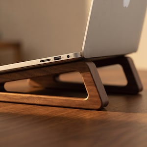 Può includere: Un computer portatile argentato poggiato su un supporto in legno marrone scuro. Il supporto presenta un design geometrico a telaio aperto con supporti angolati che sollevano il computer. Il profilo laterale mostra una porta USB e un jack audio.