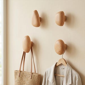 May include: Four light-colored wooden wall hooks with smooth, rounded, pebble-like shapes. Two hooks are mounted empty on the wall, while the bottom two hooks hold a woven straw tote bag and a light-colored jacket on a wooden hanger respectively.