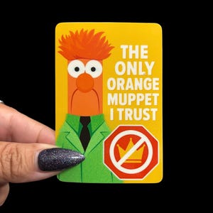 May include: A yellow rectangular sticker featuring an orange Muppet character with the text "THE ONLY ORANGE MUPPET I TRUST." The character wears a green jacket and a black tie. A red and white stop sign with a crossed-out crown is also present.