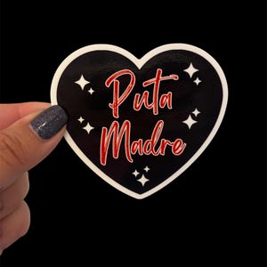 May include: A heart-shaped sticker with a black background and white border. The sticker features the words "Puta Madre" in red script, surrounded by white starbursts.