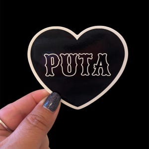 May include: A black heart-shaped sticker with a white border. The word "PUTA" is printed in white, stylized lettering across the center of the heart. The sticker is held against a black background.