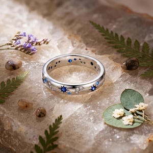 May include: A silver band ring featuring a celestial motif with inlaid blue stones, clear crystals, and engraved star and crescent moon shapes. The ring has a polished metallic finish and a repeating pattern of stars and moons around the entire band.