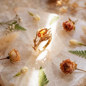 May include: A gold ring featuring a marquise-cut amber-colored gemstone set within a detailed metal band shaped like a blooming flower bud with textured leaves and stems, showcasing intricate engravings and a polished metallic finish.