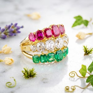 May include: A gold multi-row ring featuring three distinct bands of gemstones. The top row contains five vibrant red rubies, the middle row features five clear white diamonds, and the bottom row displays five deep green emeralds in a prong setting.