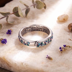 May include: A silver-toned metal ring featuring an open-ended design with a floral pattern in shades of blue, green, and pink enamel. The inner surface of the band is engraved with a lotus flower motif and several lines of Chinese characters.