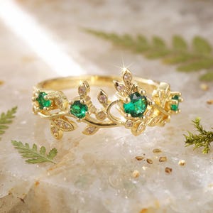 May include: A gold ring featuring a vine-inspired band with multiple small, clear, round-cut stones and four prominent, round-cut, vibrant green stones set along the top, creating a delicate, nature-themed design with a polished finish.