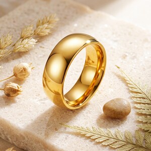 May include: A polished gold-tone band ring with a smooth, rounded exterior surface and a reflective finish. The ring is positioned upright, showcasing its circular shape and consistent width, highlighting the metallic luster and clean design.