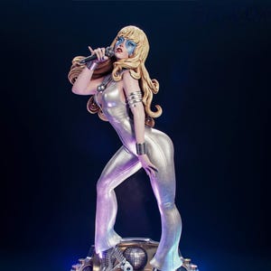 Peut inclure: Une figurine blonde portant une combinaison argentée métallisée, du maquillage bleu et des patins à roulettes argentés. Elle tient un microphone et se tient sur une base circulaire avec une boule à facettes, des détails métalliques et des lumières.