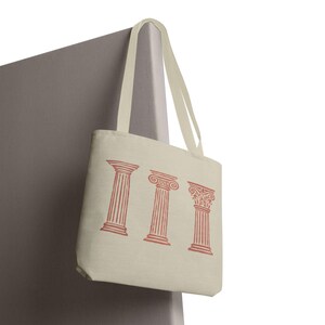 May include: A cream-colored canvas tote bag featuring a printed design of three distinct classical architectural columns in a muted terracotta red color. The bag includes two long fabric shoulder straps attached to the top edge of the rectangular body.