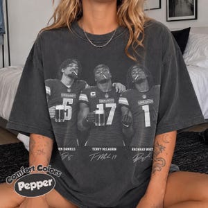 Pode incluir: Uma t-shirt cinzento-carvão oversize com um gráfico a preto e branco de três jogadores de futebol americano com camisolas numeradas 5, 17 e 1, cada um segurando uma caneca de cerveja. O texto inclui nomes, a equipa Commanders e assinaturas.