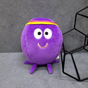 May include: A round, purple plush toy with a soft texture, featuring two large white eyes with black pupils, a stitched pink smile, and a horizontal yellow and pink headband across the top. The character has four small, stubby purple legs at the base.