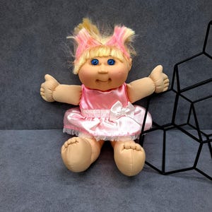 Könnte beinhalten: Eine Puppe mit blondem Haar mit rosa Strähnen, blauen Augen und einem runden Gesicht. Die Puppe trägt ein glänzendes rosa ärmelloses Kleid mit einer weißen Schleife an der Taille und einem weißen Spitzenbesatz am Saum. Helle Haut und weiche Gliedmaßen.