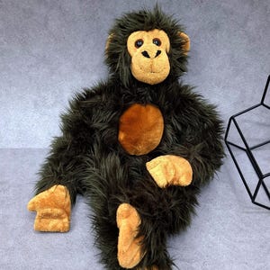 Peut inclure: Une peluche singe avec une longue fourrure synthétique vert olive foncé, un visage, des mains et des pieds beige clair, ainsi qu'une tache circulaire beige sur la poitrine. Le jouet a des yeux marron foncé et un museau brodé.