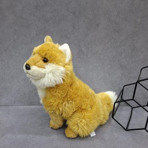 May include: A plush toy shaped like a fox with golden-brown fur, a white chest, white inner ears, and a white-tipped tail. The stuffed animal has a black nose and dark eyes, and it is positioned next to a black geometric wire frame structure.