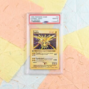 May include: A 1999 Pokemon Game 1st Edition Zapdos holographic trading card graded GEM MT 10, encased in a clear plastic PSA slab. The card features the yellow electric bird character, 90 HP, and Thunder and Thunderbolt attack text.