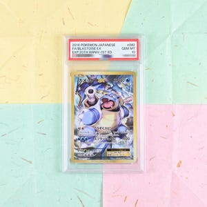 May include: A 2016 Japanese Pokemon Blastoise EX trading card, graded Gem Mint 10, encased in a clear protective plastic slab. The card features a blue Blastoise character with cannons, Japanese text, and HP180. The label reads 2016 POKEMON JAPANESE FA/BLASTOISE EX EXP.20TH ANNIV-1ST ED. #092 GEM MT 10.