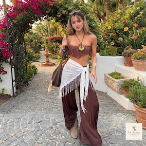 May include: A brown strapless fringe top, wide-leg brown trousers, and a white chevron-patterned sarong with fringe trim. The outfit includes a long necklace with a circular pendant, a brown shoulder bag, and tan ankle boots.