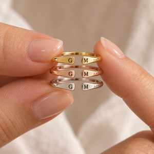 Könnte beinhalten: Drei offene Ringe, die vertikal übereinander gestapelt sind. Jeder Ring trägt die eingravierten Buchstaben G und M an den flachen, abgerundeten Enden. Der oberste Ring ist goldfarben, der mittlere roségoldfarben und der untere silberfarben.