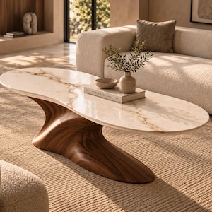 Puede incluir: Una mesa de centro ovalada con un tablero de mármol blanco con vetas marrones y una base escultural de madera maciza con una forma orgánica fluida. La superficie de la mesa sostiene un pequeño cuenco de cerámica, un libro titulado KIN FOLK y un jarrón.