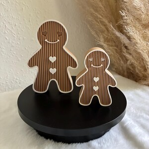 Könnte beinhalten: Zwei lebkuchenmannförmige Figuren in Braun mit vertikalen schwarzen Streifen und weißen Umrandungen. Jede Figur hat ein lächelndes Gesicht mit herzförmigen Augen und zwei weiße herzförmige Knöpfe auf der Brust. Sie stehen auf einem schwarzen Sockel.