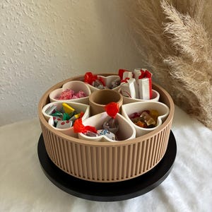Könnte beinhalten: Ein rundes, beiges, geripptes, drehbares Snack-Tablett mit sechs weißen, herzförmigen Fächern, die um einen zentralen Zylinder angeordnet sind. Die Fächer sind mit verschiedenen einzeln verpackten Bonbons und bunten Fruchtgummis gefüllt.