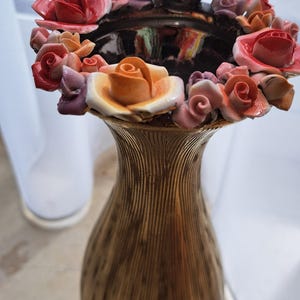 May include: A tall, slender vase with a textured, metallic gold finish and vertical ribbed detailing. The rim is adorned with a dense arrangement of small, three-dimensional ceramic roses in shades of pink, orange, peach, and deep purple.