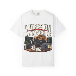 May include: A white t-shirt featuring a graphic of a dog wearing sunglasses, sitting at a desk with two computer monitors, a laptop, a mug, and a stack of books. The text "WORKS ON MY MACHINE" is printed in large letters above the illustration.