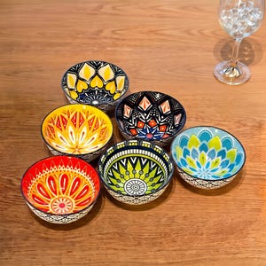 Può includere: Un set di sei piccole ciotole in ceramica con intricati e colorati motivi in stile mandala nelle tonalità del giallo, arancione, rosso, verde, blu e nero. Ogni ciotola presenta un design geometrico unico all'interno e un motivo costante in bianco e nero sul bordo esterno.