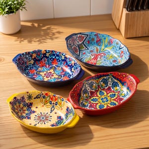 May include: Four oval ceramic baking dishes with handles, each featuring a distinct, vibrant, multi-coloured floral and geometric pattern. The dishes are coloured in shades of blue, red, and yellow, showcasing intricate, symmetrical designs.