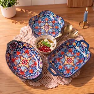 May include: A set of three ceramic serving dishes featuring a vibrant blue, red, and yellow floral mandala pattern. The collection includes one flower-shaped plate, one oval baking dish with handles, and one rectangular baking dish with handles.