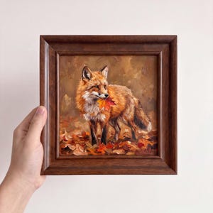 Può includere: Un dipinto a olio quadrato in una cornice di legno marrone scuro raffigurante una volpe rossa in piedi tra le foglie autunnali cadute. La volpe ha il pelo arancione e bianco e tiene in bocca una foglia d'acero rossa e arancione.