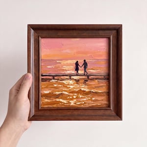 Puede incluir: Una pintura al óleo en un marco de madera marrón oscuro que muestra a dos figuras tomadas de la mano en un muelle al atardecer. La escena presenta pinceladas gruesas y texturizadas en tonos rosa, naranja y amarillo dorado sobre el agua.
