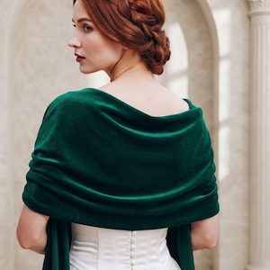 Puede incluir: Un chal de terciopelo verde esmeralda profundo drapeado sobre los hombros, con una textura suave y afelpada y un diseño ancho y envolvente que cubre la parte superior de la espalda y los brazos, sobre una prenda blanca con botones.