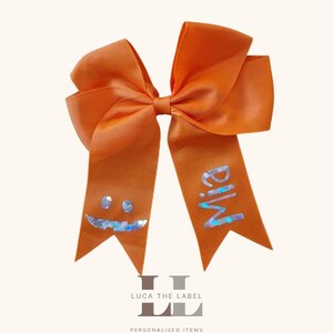 May include: A bright orange hair bow made of ribbed fabric with two long tails. One tail features a holographic smiley face graphic, and the other tail displays the name Mia in holographic lettering. The bow has a classic multi-loop design.