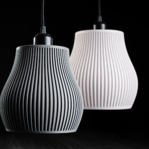 May include: Two pendant lights with bell-shaped shades featuring vertical ribbed textures. One shade is matte gray and the other is matte white. Each light is suspended by a black cord attached to a black socket at the top of the fixture.
