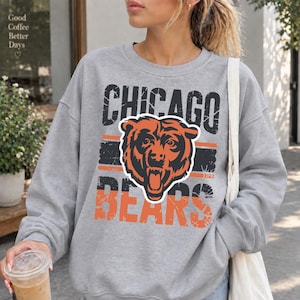 May include: A heather gray crewneck sweatshirt featuring a distressed graphic print on the front. The design includes the text CHICAGO BEARS in black and orange, centered around a stylized illustration of a roaring bear head in orange and black.